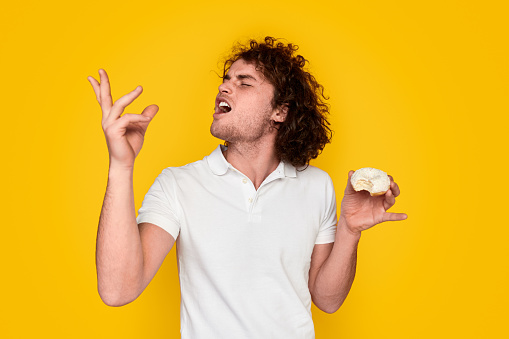 Young male with curly hair keeping eyes closed and gesturing with hand while tasting marvelous donut against vibrant yellow background