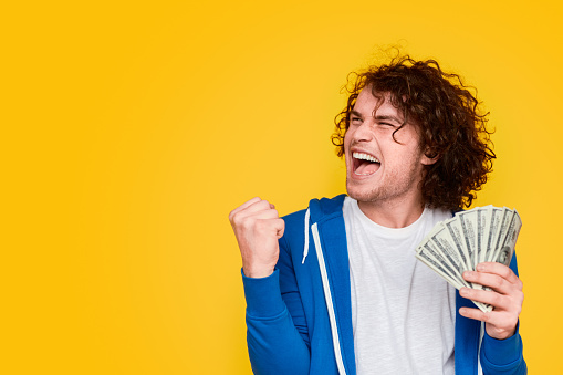 Cheerful young man with bunch of money clenching fist and looking at empty space after winning lottery against yellow background