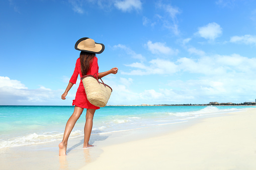 Beachwear woman tourist with straw sun hat and beach bag walking on tropical summer vacation wearing sunhat and red tunic dress cover-up relaxing on travel holidays from behind.