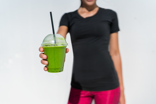 Healthy eating lifestyle fitness athlete woman drinking green smoothie cup for vegan weight loss diet. breakfast juicing trend. Person showing plastic container at outdoor cafe or juice bar.