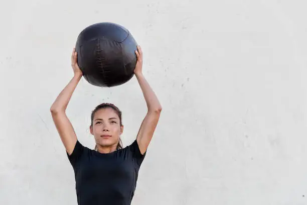 Fitness girl holding medicine ball above head for shoulder press workout in outdoor gym. Young Asian athlete girl doing upper body exercise working out with heavy weighted balls.