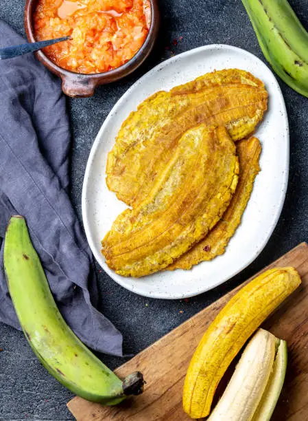 Photo of COLOMBIAN CARIBBEAN CENTRAL AMERICAN FOOD. Patacon or toston, fried and flattened whole green plantain banana on white plate with tomato sauce and chicharron Black background, top view