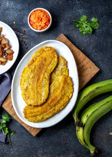 Photo of COLOMBIAN CARIBBEAN CENTRAL AMERICAN FOOD. Patacon or toston, fried and flattened whole green plantain banana on white plate with tomato sauce and chicharron Black background, top view