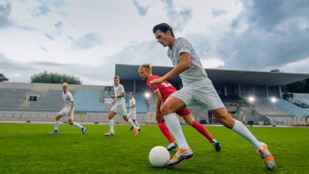 Professional Soccer Player Leads with a Ball, Masterfully Dribbling and Bypassing Sliding Tackles of His Opponents. Two Professional Football Teams Playing. Low Angle Shot. Professional Soccer Player Leads with a Ball, Masterfully Dribbling and Bypassing Sliding Tackles of His Opponents. Two Professional Football Teams Playing. Low Angle Shot. defending sport stock pictures, royalty-free photos & images