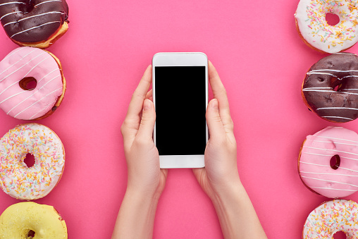 cropped view of woman holding smartphone with blank screen near tasty glazed doughnuts on bright pink background