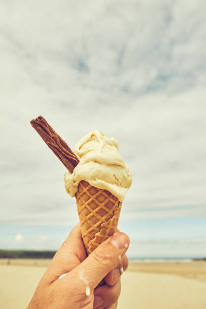 istock POV hand holding a Vanilla Ice cream cone at Crantock Beach, Newquay, Cornwallmon a June day, mild vintage film tone. 1161551525