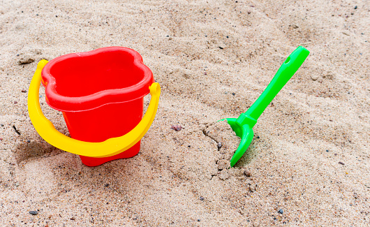 This is a close up photo of a children's colorful striped beachball next to a plastic toy bucket shovel and rake sitting on sand and photographed in the studio isolated on a white background.