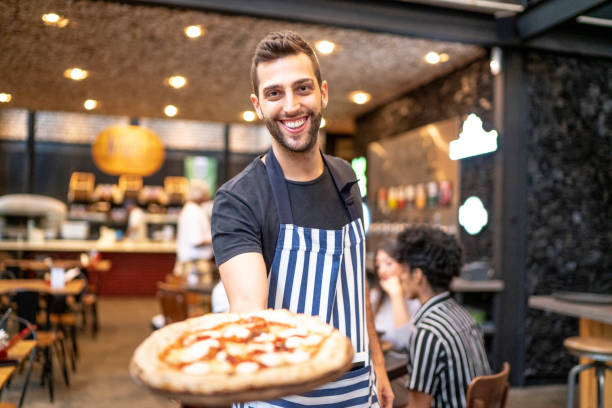 cameriere sorridente che guarda la macchina fotografica e mostra una pizza - pizzeria foto e immagini stock