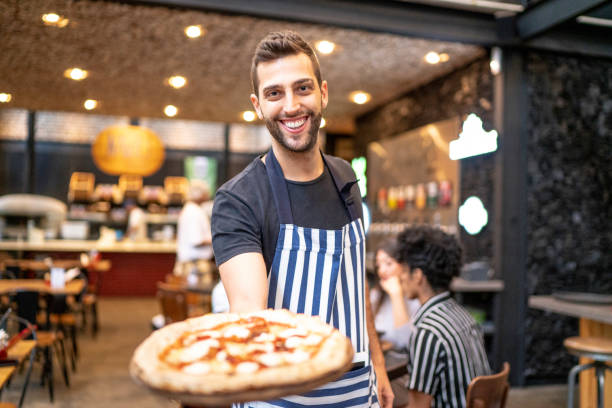 smiling waiter looking at camera and showing a pizza - pizzeria imagens e fotografias de stock