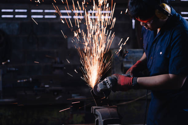 Immigrant manufacturing worker using angle grinder power tools on metal creating hot sparks wearing safety protective work wear stock photo