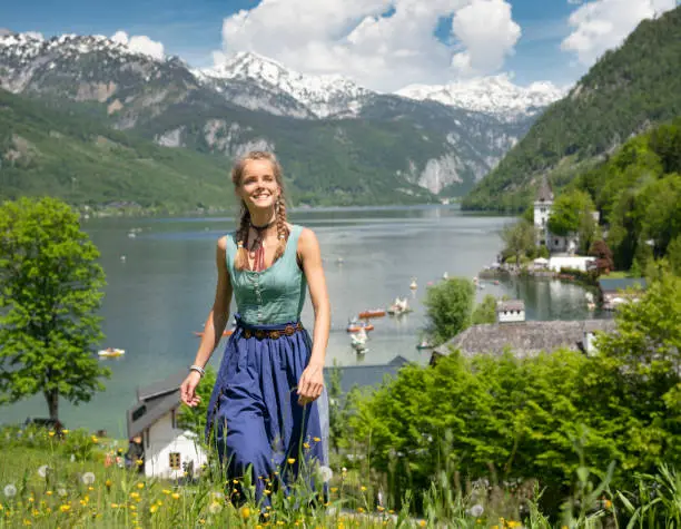 Natural Beauty wearing a traditional Dirndl walking up a meadow full of flowers in front of famous Grundlsee, Austrian Alps Panorama. You can see the Boat Corso of the popular Narzissenfest on the Lake in back. Grundlsee, Steiermark, Ausseerland, Austria. Nikon D850. Converted from RAW.