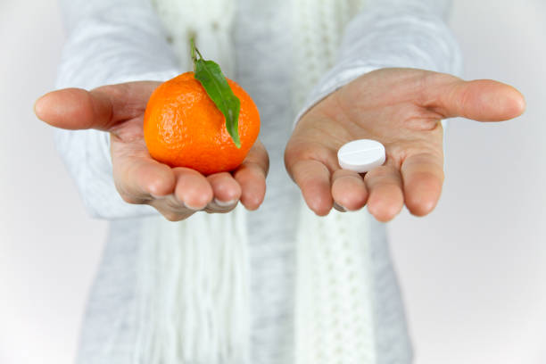 vitamins from fruit or drugs? a sick young woman with a scarf on her neck shows a mandarin in her right hand and an aspirin on her left - vibrant color healthcare and medicine healthy lifestyle vitamin pill imagens e fotografias de stock