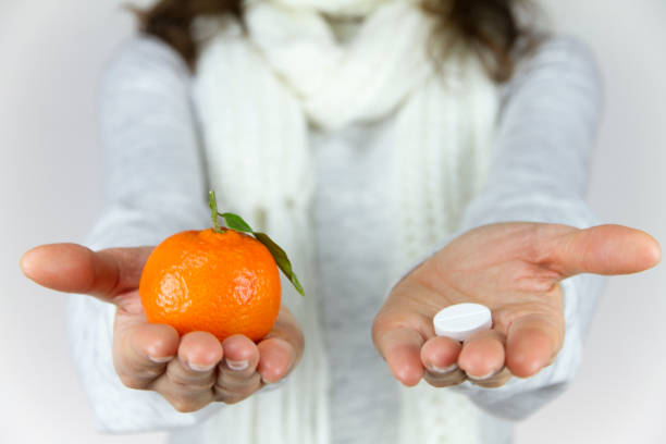 vitamins from fruit or drugs? a sick young woman with a scarf on her neck shows a mandarin in her right hand and an aspirin on her left - vibrant color healthcare and medicine healthy lifestyle vitamin pill imagens e fotografias de stock