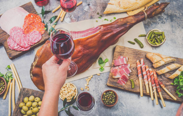 mans hand holding wine glass frome above table with spanit or italian appetizers, whole leg of ham serrano and red an rose wine - serrano chilli pepper meat ham spain imagens e fotografias de stock