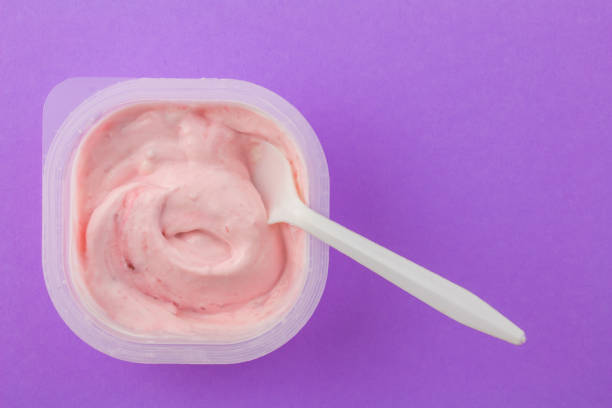 istock Yogurt cup with strawberry yogurt and with white plastic spoon dipped isolated on purple background 1161586785