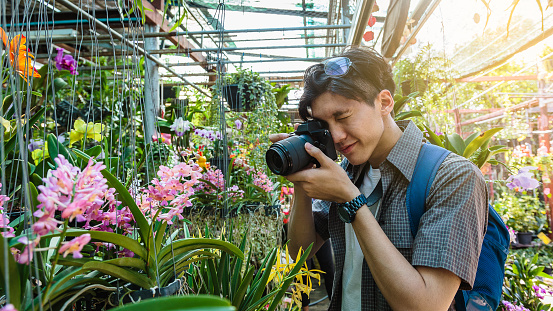 Young Asian traveller man taking pictures with camera to orchid and other plants at traditional street market, Khonkaen city, Thailand