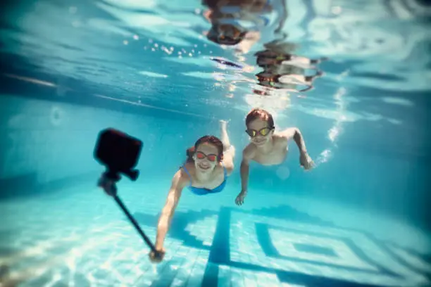 Brothers and sister are having fun playing underwater in the resort pool.  Kids are filming themselves using waterproof action camera on selfie stick.
Nikon D810