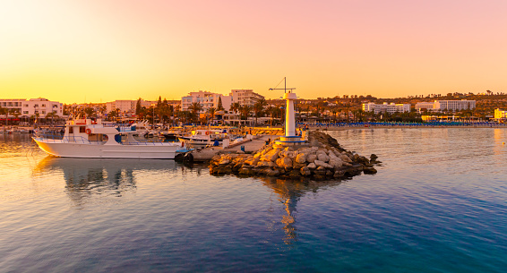 Ayia Napa, Cyprus - May 01, 2018: Ayia Napa harbor and lighthouse. Tourist boats and fishing boats. Sunset time. People on the background.