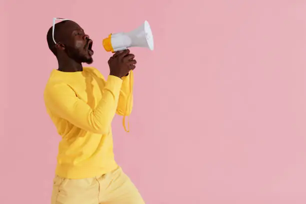 Black man shouting in megaphone on pink background portrait. Happy young male model screaming in horn speaker in studio