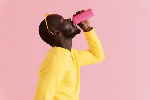 Drink. Black man drinking soft drink on pink background. Colorful portrait of happy smiling african american male model in yellow fashion clothes enjoying soda from pink can in studio