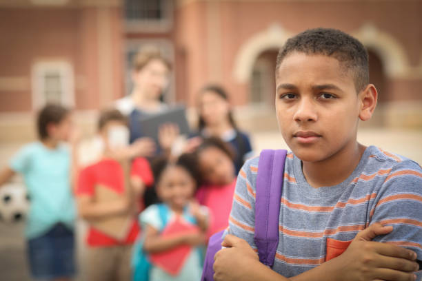 Junior high age boy being bullied at school. Sad, junior high school age boy being bullied outside the school building,  A group of multi-ethnic students in background laugh at the African American boy.  They have mobile devices to cyber bully the child as well as point and talk about him. sad child standing stock pictures, royalty-free photos & images