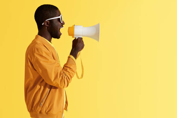 Advertising. Man screaming announcement in megaphone portrait Advertising. Man screaming announcement in megaphone on yellow background. Portrait of african american male model in fashion wear using loud speaker in studio black orange audio stock pictures, royalty-free photos & images