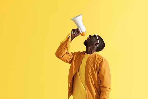 Advertising. Man screaming announcement in megaphone on yellow background. Portrait of african american male model in fashion wear using loud speaker in studio