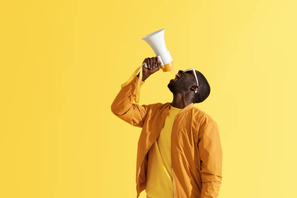 Advertising. Man screaming announcement in megaphone portrait Advertising. Man screaming announcement in megaphone on yellow background. Portrait of african american male model in fashion wear using loud speaker in studio toned image stock pictures, royalty-free photos & images