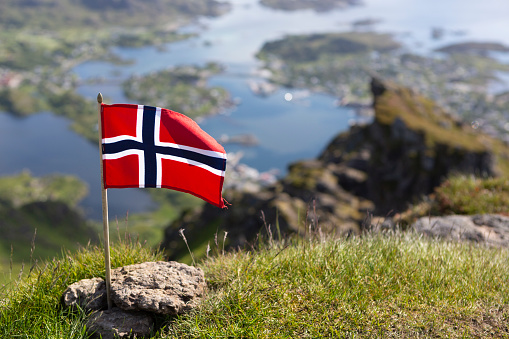 Norwegian Flag on the Nonstinden mountain. Blurred in the background the view down to Ballstad, Vestvågøya island, Lofoten Islands, Norway