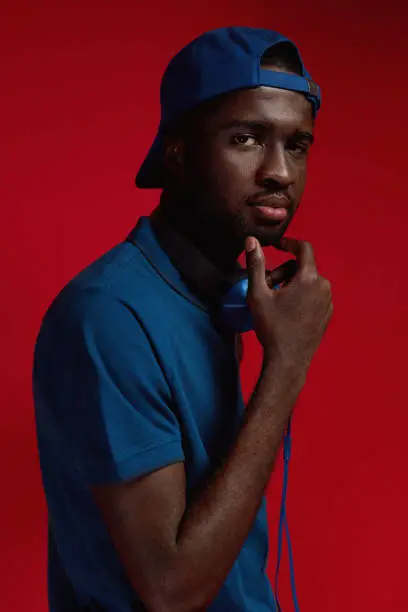 Fashion portrait of black man in stylish outfit with headphones at studio. African male model in blue cap, polo shirt and headphone on red background