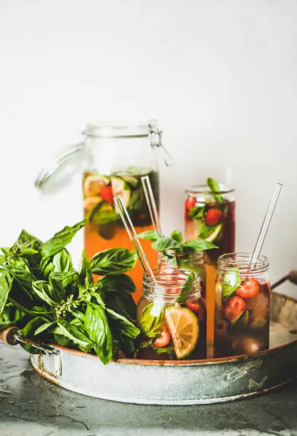 Fresh homemade strawberry and basil lemonade or iced-tea in glass tumblers with eco-friendly glass plastic-free straws on rustic metal tray, white wall background behind. Summer cold refreshing soft drink