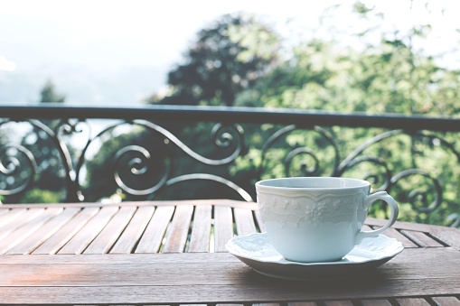 Single white ceramic cup of tea on a saucer on wooden table against picturesque and natural green foliage background.