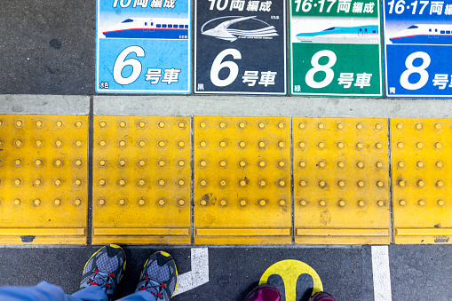 Utsunomiya, Japan - April 4, 2019: Looking down high angle view of couple woman and man people legs or feet standing on platform with train station car numbers by yellow line