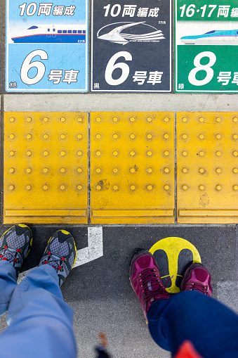 Utsunomiya, Japan - April 4, 2019: Looking down high angle view of couple, people legs or feet standing on platform with train station car numbers by yellow line