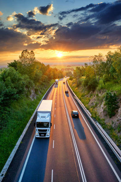 Three white truck and three private car driving on the asphalt road in a rural landscape at sunset with dramatic clouds. View from above. Three white truck and three private car driving on the asphalt road in a rural landscape at sunset with dramatic clouds. View from above. semi auto stock pictures, royalty-free photos & images