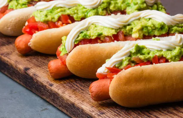 Photo of Chilean Completo Italiano. Hot dog sandwiches with tomato, avocado and mayonnaise on wooden board. closeup