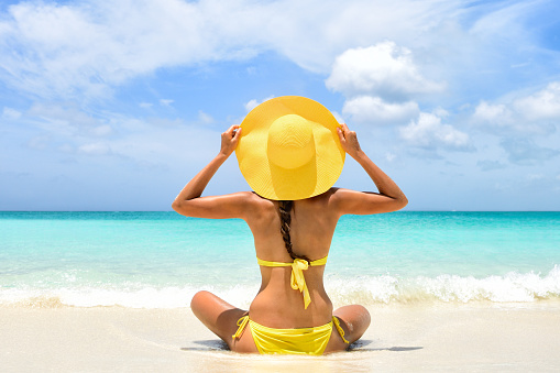 Happy carefree woman relaxing sitting in sand enjoying tropical beach destination. Perfect paradise summer vacation happiness. Back view of bikini girl holding yellow fashion hat on Caribbean holiday.