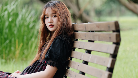 Portrait of beautiful Asian woman sitting on bench in summer forest, Chinese girl in vintage black dress looking at camera with blur nature background, Emotions, beauty and lifestyle concept.