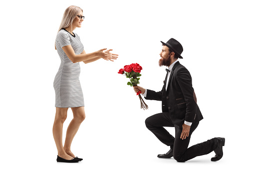 Full length profile shot of a bearded man kneeling and giving a bouquet of red roses to a woman isolated on white background