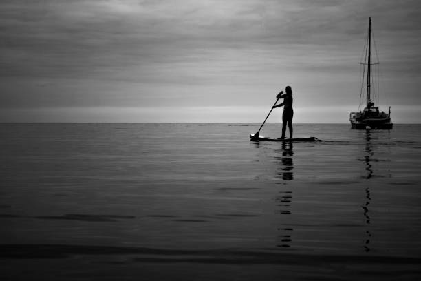 A black and white photo of a  young woman on a paddle board casting long reflections in the sea on a calm evening.