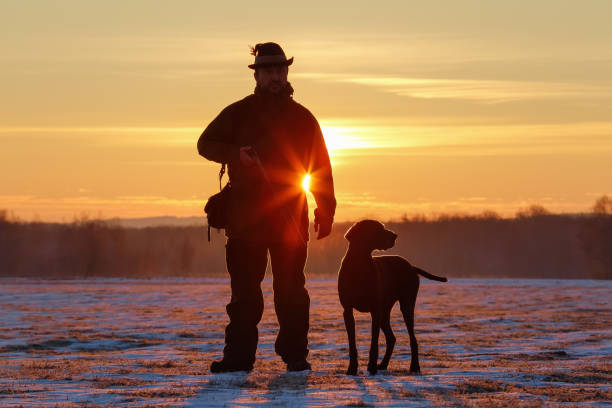 istock A hunter in the hat with a gun and his German Shorthaired Pointer dog breed friend. Beautiful silhouettes on the background of the dawn. Amazing winter morning. 1161740366
