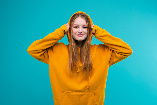 Portrait of a young emotional woman in a yellow hoodie isolated over blue background