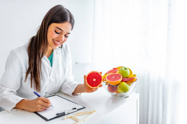 Portrait of young smiling female nutritionist in the consultation room Smiling nutritionist in her office, she is showing healthy vegetables and fruits, healthcare and diet concept. Female nutritionist with fruits working at her desk. nutritionist stock pictures, royalty-free photos & images