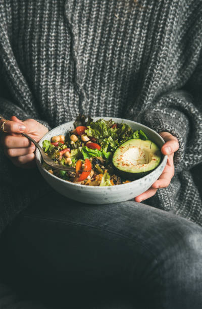 Woman in sweater eating healthy vegetarian dinner from Buddha bowl Healthy vegetarian dinner. Woman in grey jeans and warm sweater holding bowl with fresh salad, avocado, grains, beans, roasted vegetables. Superfood, clean eating, vegan, dieting food concept foxys_forest_manufacture stock pictures, royalty-free photos & images