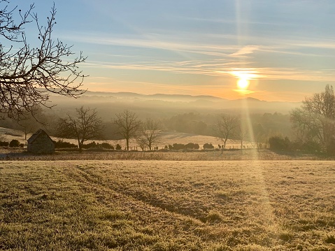 Sunrise on a crisp winter morning in the French countryside, hoarfrost, sunbeams coming through the mist, horizontal view taken from hill, poetic nature background