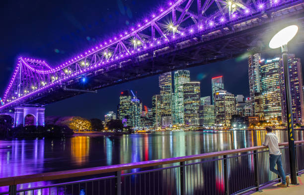 View of bright bridge in purple lights above river of city with embankment and skyscrapers, Brisbane