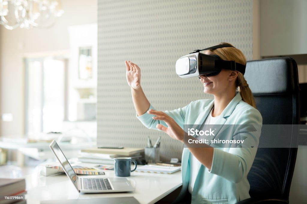 Young female employee in vr goggle attenting virtual training Young female employee in vr goggle attenting virtual training while sitting by desk in office Virtual Reality Simulator Stock Photo
