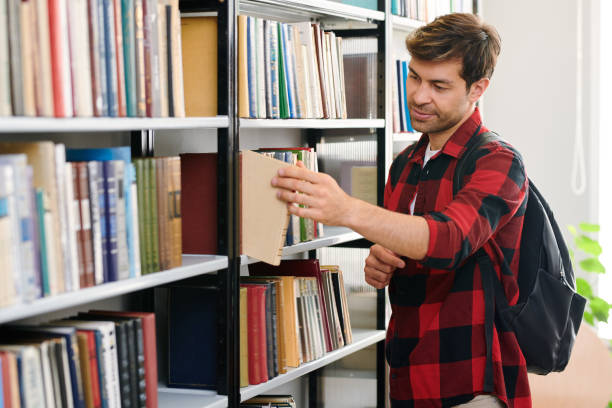 istock Young student with backpack taking one of books from shelf in library 1161792586