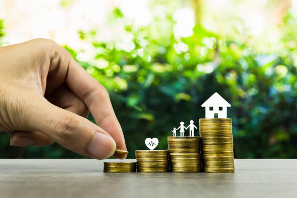 Saving or making money for family life in future concept. A man hand putting coin into rising stack of coins with heart, family and small house model on wood table. Depicts sustainable financial goal. ahorrar o ganar dinero para la vida familiar en el concepto futuro. un hombre a mano poniendo moneda en la pila creciente de monedas con corazón, familia y modelo de casa pequeña en la mesa de madera. representa un objetivo financiero sostenible.
