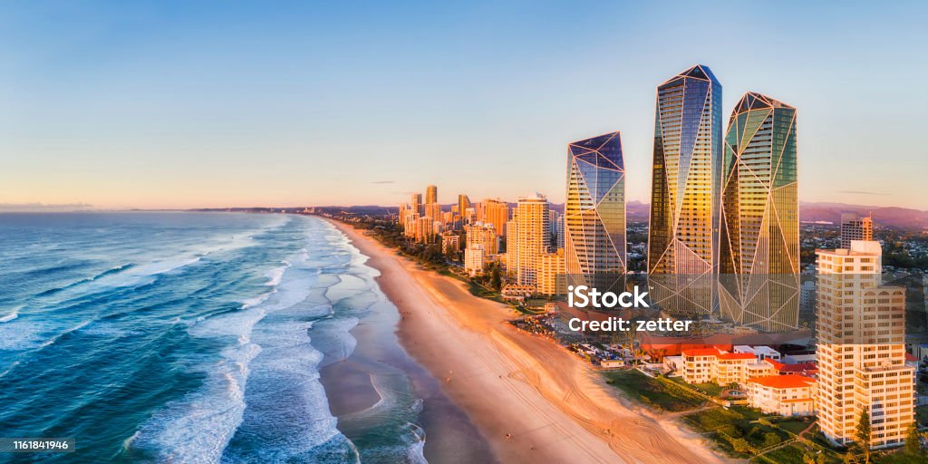 D QLD SP rise 2 south pan Rising sun shining on modern urban towers of Surfers paradise in Australian Gold Coast facing endless waves of Pacific ocean - aerial panoramic view. Australia Stock Photo